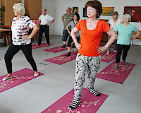 Auf dem Boden eines Saals liegen violette Yogamatten. Auf jeder Yogamatte steht eine Kursteilnehmerin oder ein Kursteilnehmer. Sie haben das linke Bein nach vorn gestellt, stehen aufrecht, die Hände in die Taille gestützt. Es sind Menschen im mittleren und höheren Alter, vorwiegend Frauen.