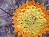 Eine Teil einer Blüte aus Keramik mit violetten Blütenblättern und einem gelben Kreis aus Staubblättern, welcher von einem orangen schmalen orange-farbenen Ring kleiner Blättchen umgeben ist